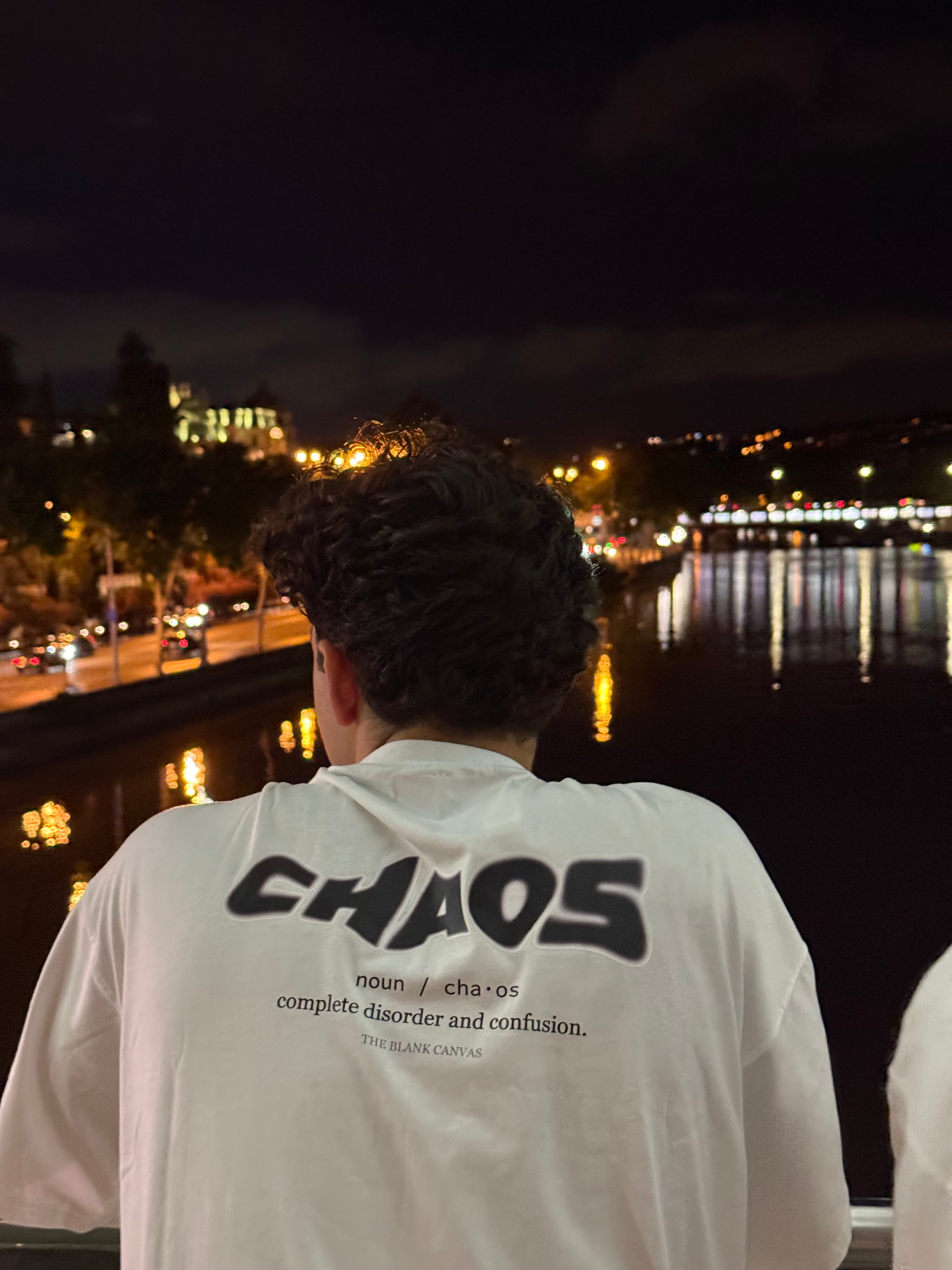Person wearing a white shirt with 'CHAOS' text, standing against a night cityscape.