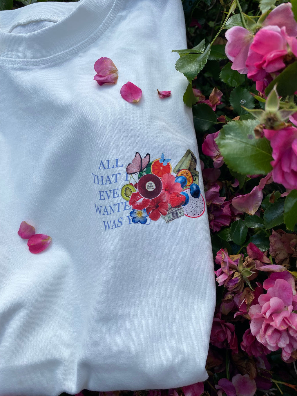 White shirt with floral design and text, surrounded by pink flowers and green leaves.