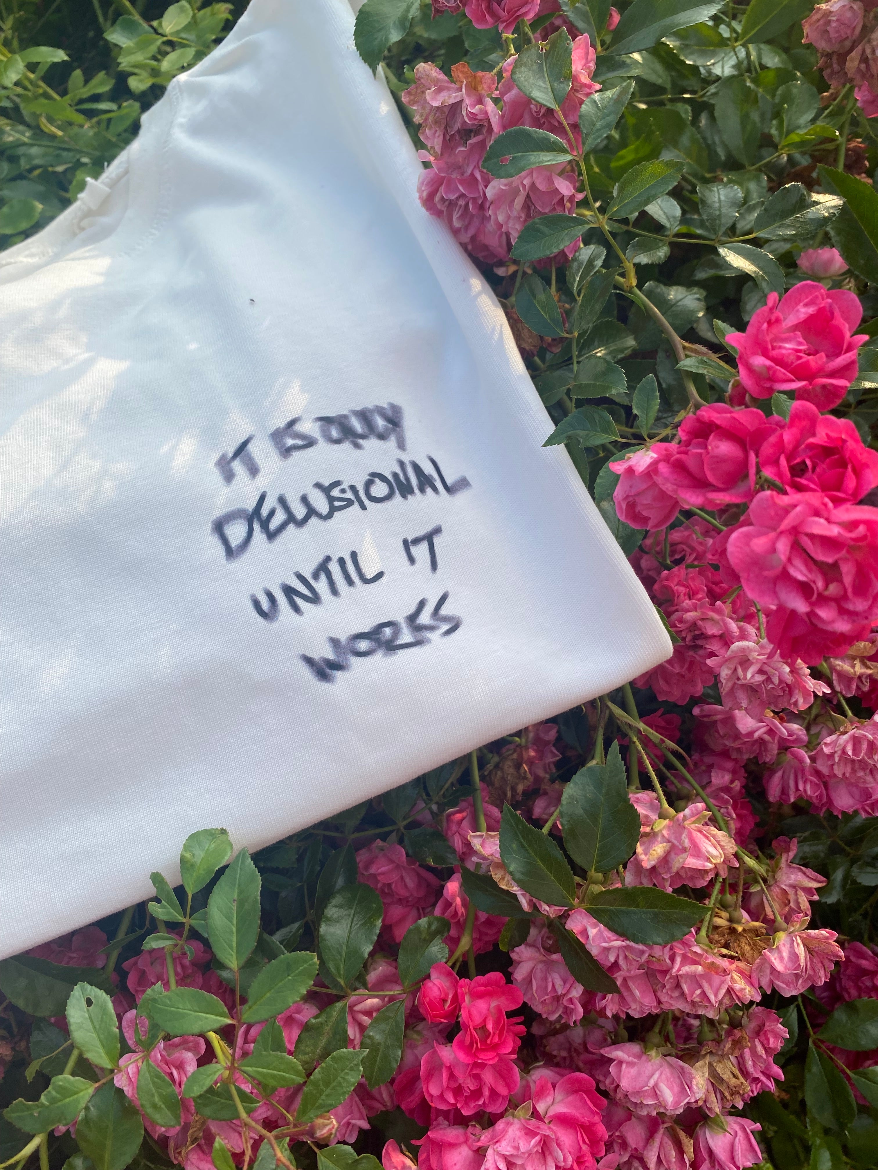 White shirt with text displaying "Its only Delusional Until It Works" on a background of pink flowers