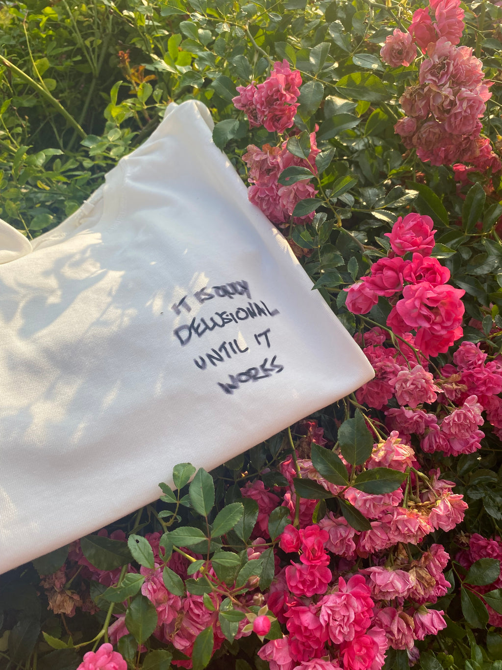 White shirt with text displaying "Its only Delusional Until It Works" on a background of pink flowers